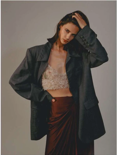 A woman in an oversized blazer poses in front of a neutral background, showcasing a textured top and maroon-colored skirt.
