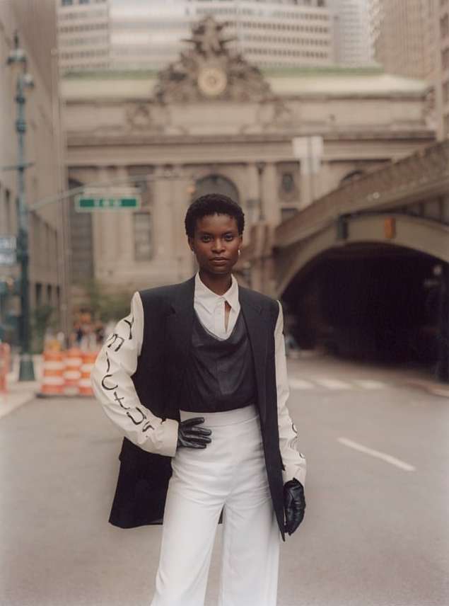 Fashion shot of a Black woman in black and white business-casual attire, striking a pose on a city street with buildings in background.