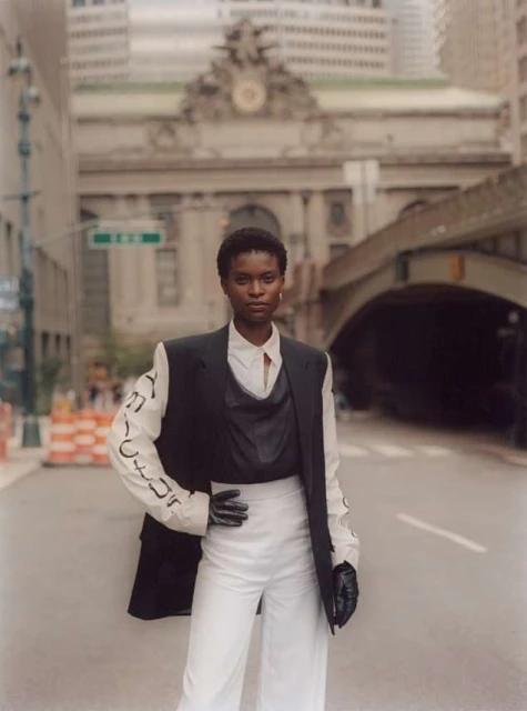 Fashion shot of a Black woman in black and white business-casual attire, striking a pose on a city street with buildings in background.