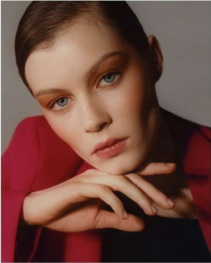 Close-up portrait of a young woman with light skin, blue eyes, and auburn hair, wearing a red blazer in a photo studio setting.