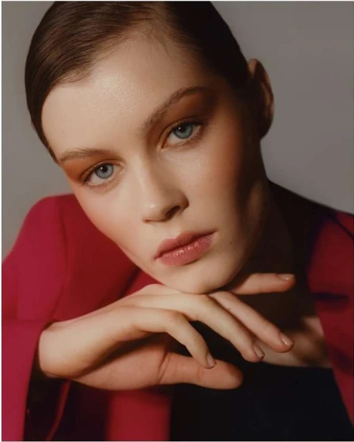 Close-up portrait of a young woman with light skin, blue eyes, and auburn hair, wearing a red blazer in a photo studio setting.