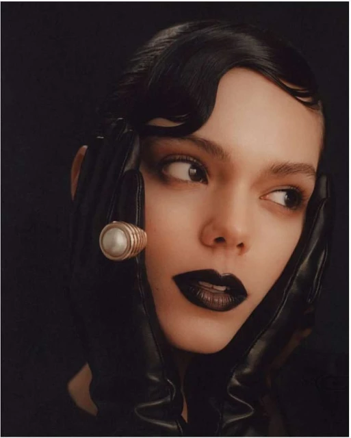 Close-up studio shot of a woman with black lipstick and slicked-back hair wearing black gloves and a large ring on her finger.