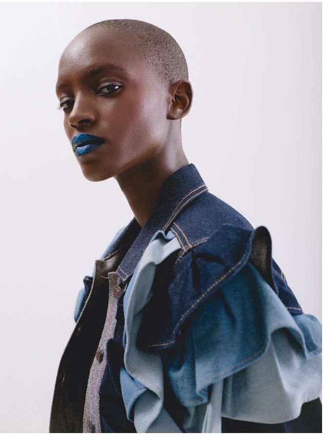 Portrait of a young Black woman with short hair wearing a denim jacket with ruffled sleeves and blue lipstick, set against a white background.