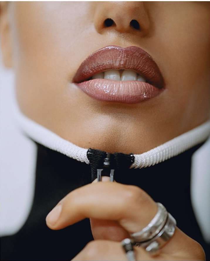 Close-up of a person's face featuring glossy lips with neutral lipstick. A hand pulls the zipper of a white and black turtleneck.