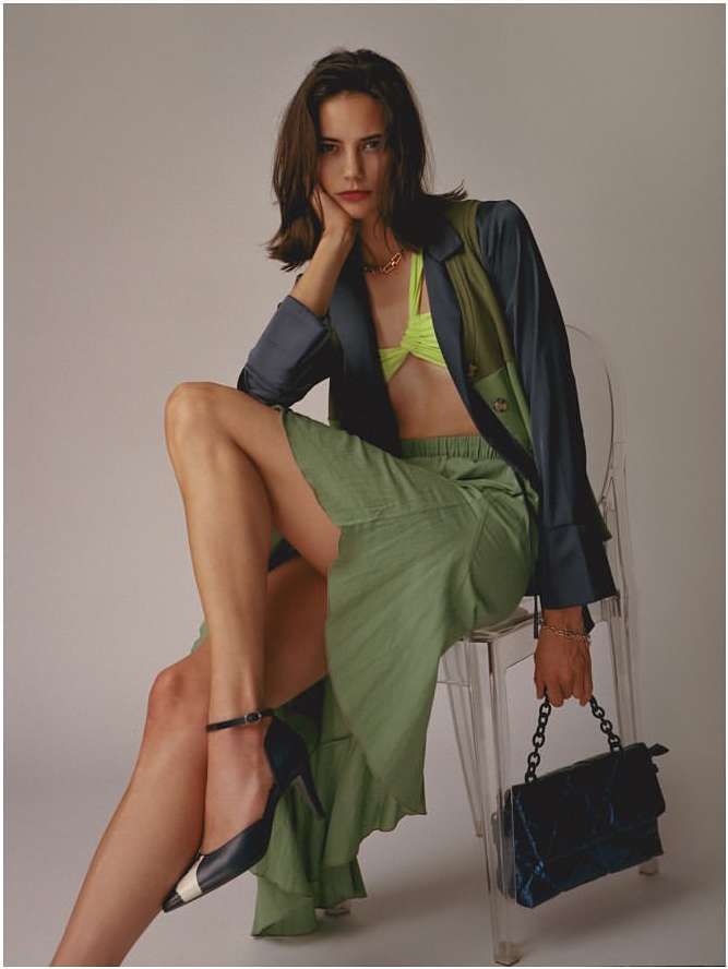 Fashionable woman with dark hair poses while seated on a clear chair in a studio, wearing a two-toned blazer, skirt, and heels.