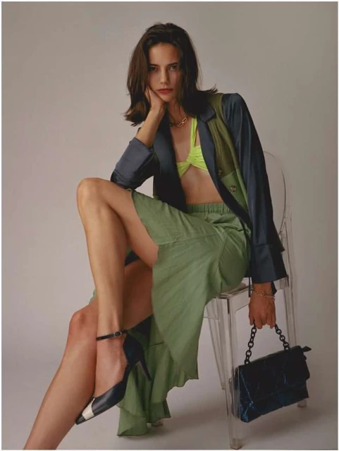 Fashionable woman with dark hair poses while seated on a clear chair in a studio, wearing a two-toned blazer, skirt, and heels.