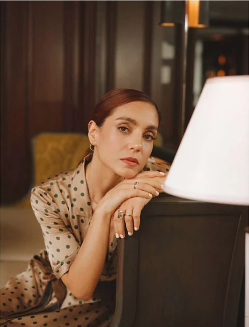 Portrait of a brunette woman in a polka dot silk dress, resting her arms on a black desk with a white lamp, indoors.
