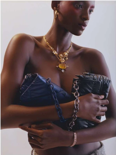 A topless Black woman poses indoors with a statement gold necklace, jewelry, and two puffy black bags with chain straps.