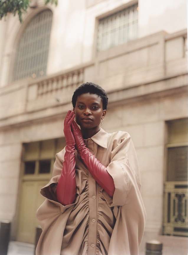 Model with short dark hair wearing long red leather gloves and a beige buttoned dress against an architectural stone building.