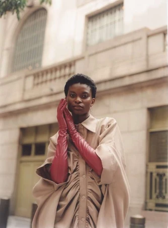 Model with short dark hair wearing long red leather gloves and a beige buttoned dress against an architectural stone building.