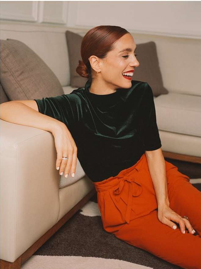 A smiling red-haired woman in a green velvet top and orange pants sits on a patterned rug near a beige couch in an indoor setting.