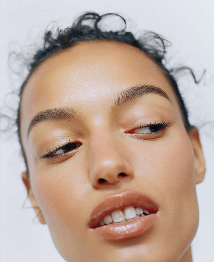 Close-up portrait of a tanned adult female with glossy lips and minimal eye makeup against a plain white background.