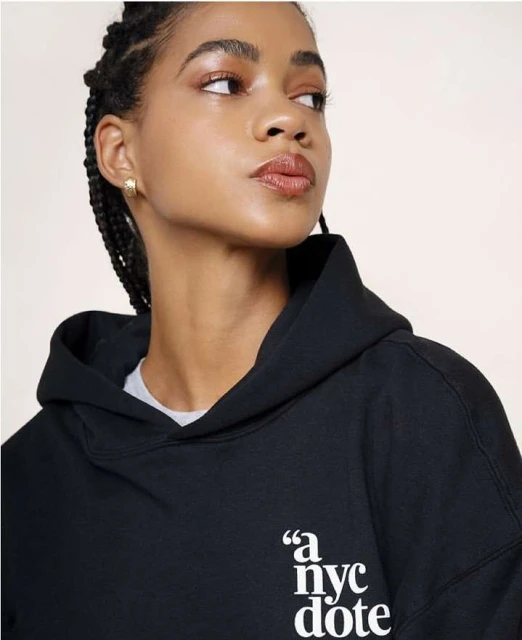 Close-up of a young adult female in a black hoodie with the words 'a nyc dote' on the front, against a bright neutral background.
