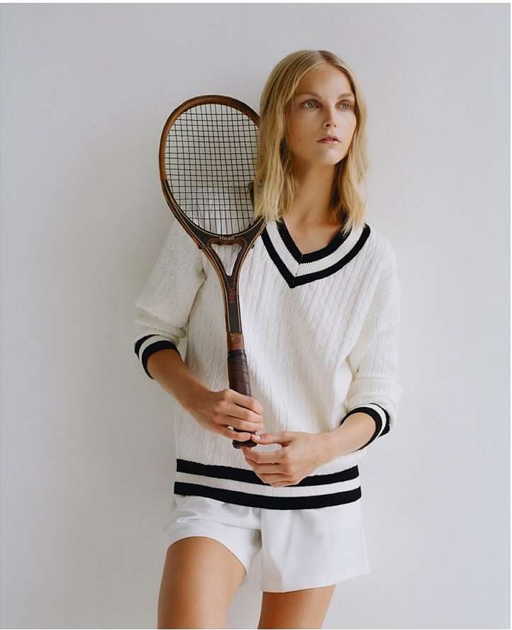 Woman in white sweater and shorts poses with vintage tennis racket in studio, looking off camera with neutral expression, soft lighting.