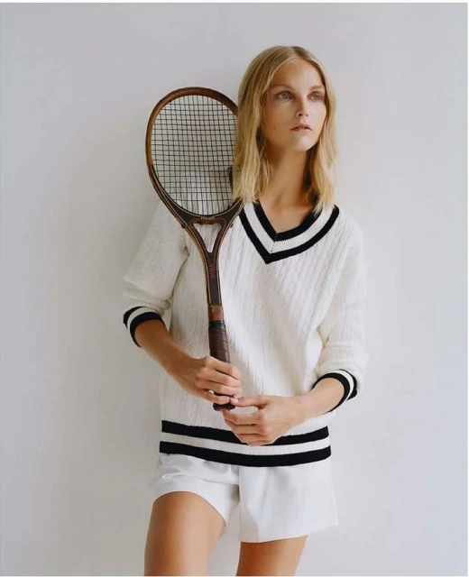 Woman in white sweater and shorts poses with vintage tennis racket in studio, looking off camera with neutral expression, soft lighting.