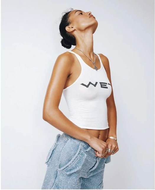 Young adult woman in a white tank top and light blue textured pants poses in a studio, looking up, with neutral facial expression.