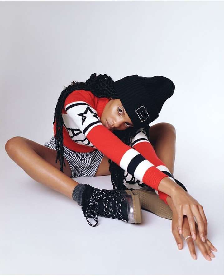 An adult female in a photo studio is angled, wearing a knit hat, a red and white shirt, and unique footwear, sitting on a white backdrop.