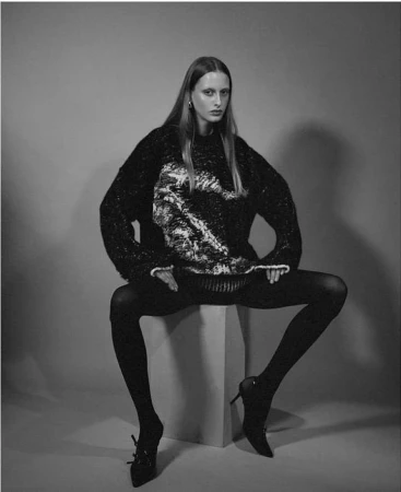 Monochrome portrait of a seated model wearing a textured dark sweater and black tights against a plain studio background.