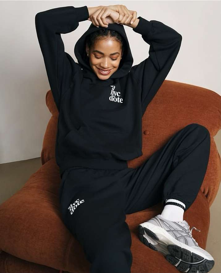 A young woman with a hoodie and sweatpants poses in a relaxed position while sitting in a chair, wearing sportswear and sneakers.