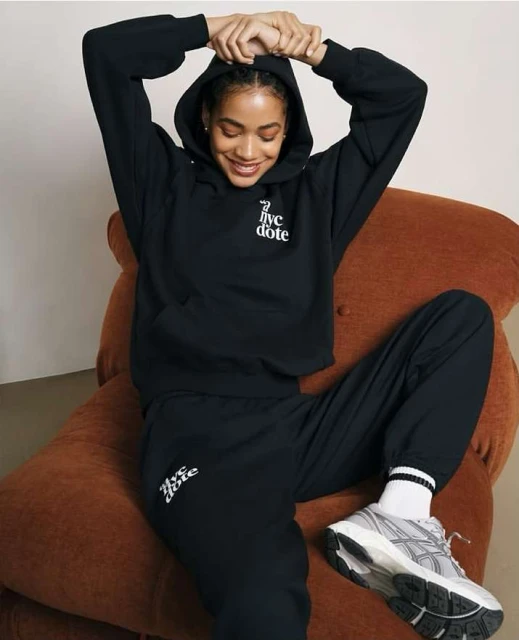 A young woman with a hoodie and sweatpants poses in a relaxed position while sitting in a chair, wearing sportswear and sneakers.