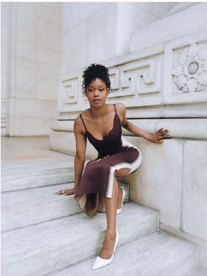 Woman with curly hair wearing a brown dress and white heels poses on marble stairs with a white architectural background indoors.