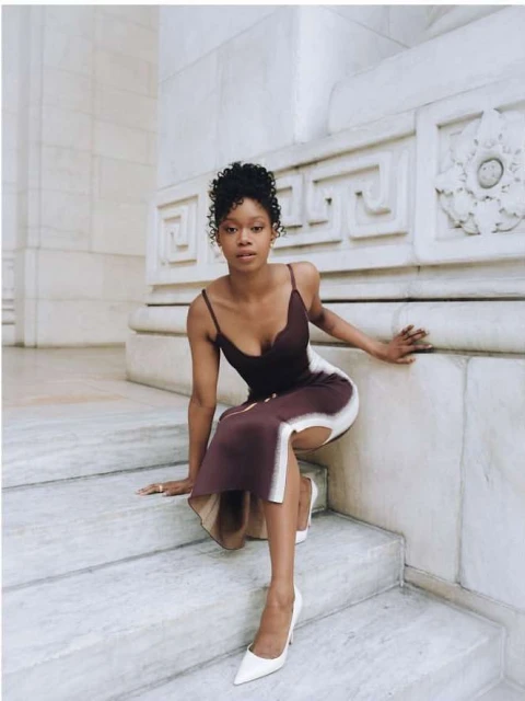 Woman with curly hair wearing a brown dress and white heels poses on marble stairs with a white architectural background indoors.