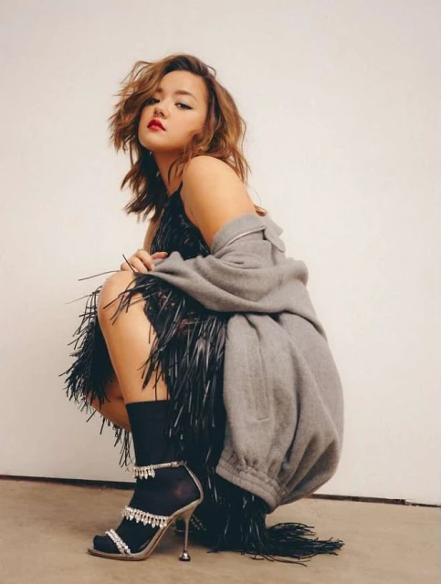Stylish woman in a black fringe dress and gray jacket poses in a studio with one leg up and wearing high-heeled sandal shoes.