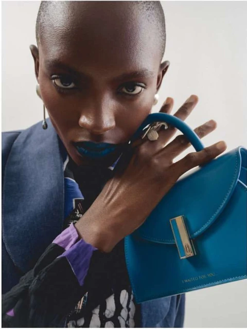 Close-up of an African American woman holding a blue leather handbag with a gold clasp, wearing a blue jacket and statement lipstick.