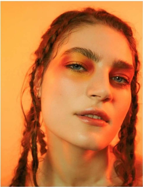 Close-up portrait of a woman with braids, featuring artistic orange and yellow eye makeup, set against a warm orange background.