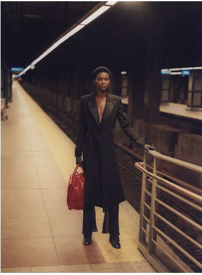 A slender woman in a black coat and pants standing in a subway station, carrying a red bag, with a neutral facial expression.