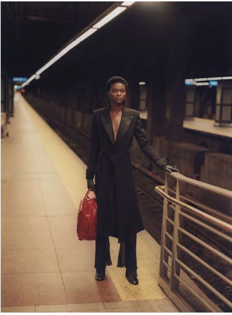 A slender woman in a black coat and pants standing in a subway station, carrying a red bag, with a neutral facial expression.