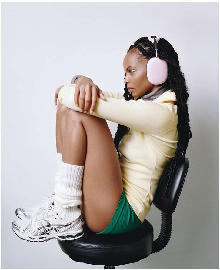 A woman sits on a black stool, wearing pink headphones, a yellow shirt, green shorts, and athletic shoes in a bright photo studio.