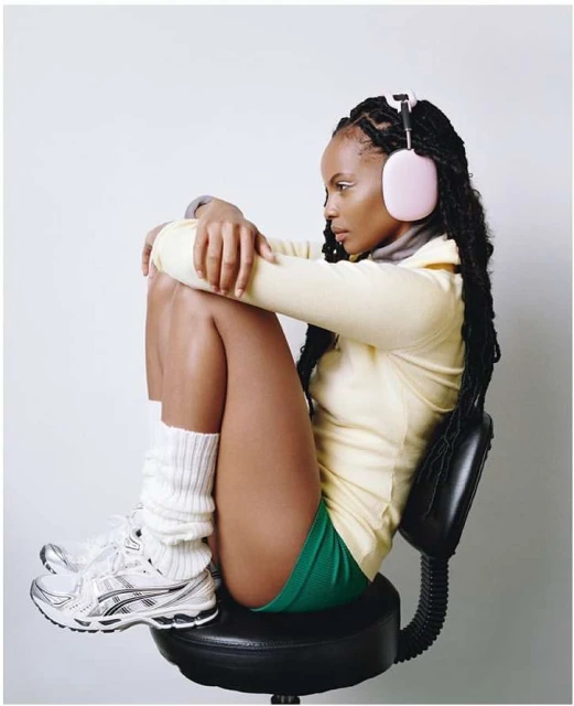 A woman sits on a black stool, wearing pink headphones, a yellow shirt, green shorts, and athletic shoes in a bright photo studio.