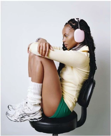 A woman sits on a black stool, wearing pink headphones, a yellow shirt, green shorts, and athletic shoes in a bright photo studio.