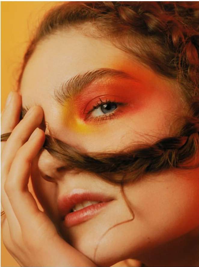 Close-up of a young woman with orange and yellow eyeshadow, styled auburn hair, and her hand near her face against an orange background.