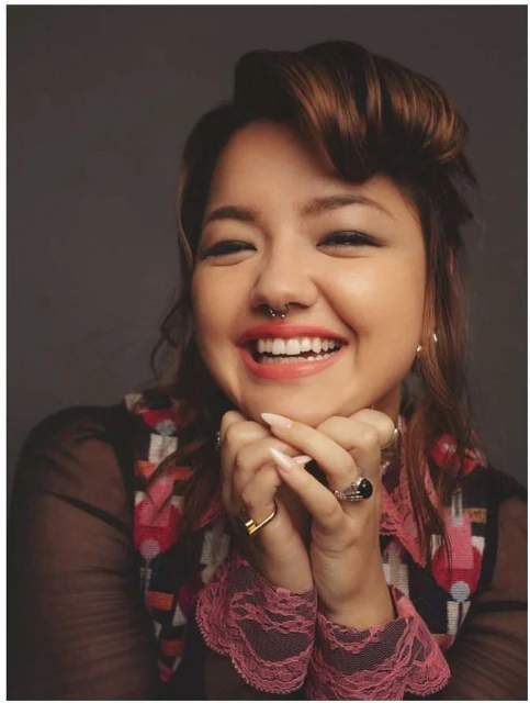 Portrait of a smiling adult woman with brown hair, wearing a sheer top and rings, resting her face on her hands against a grey backdrop.