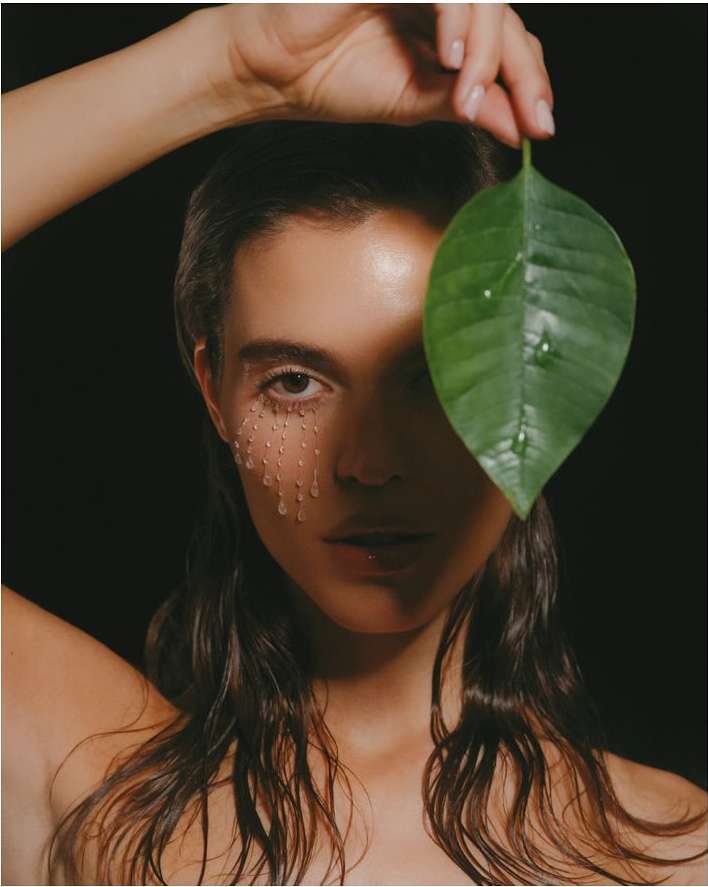 Close-up of a brunette woman with water droplets on her face, holding a large green leaf against a stark black background. Beauty shot.