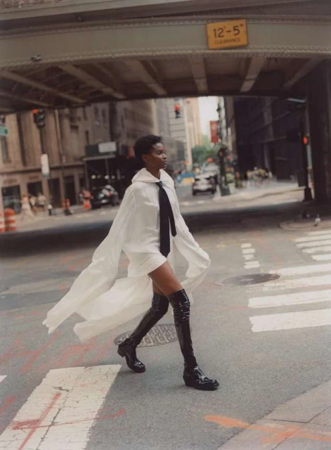 An adult female model walks across a city street in a white cape dress and shiny black knee-high boots below an overpass.
