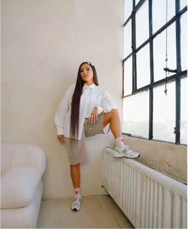 Woman with long dark hair wearing a white button-down shirt and beige shorts posing near a large multi-pane window