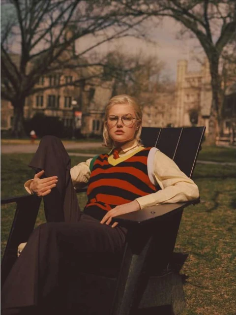 Young woman with blonde hair is sitting in a black chair outdoors, wearing glasses, a striped sweater, pants, and a collared shirt.