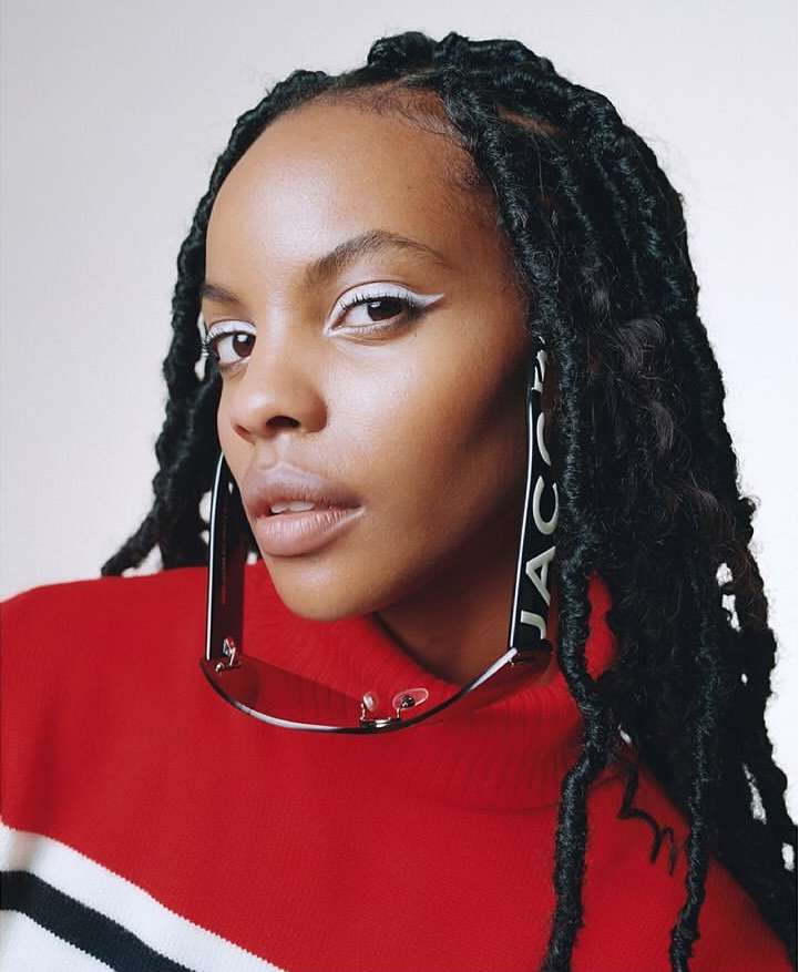 Headshot of a black woman with dreadlocks wearing a red and white sweater and sunglasses as earrings against a white background.