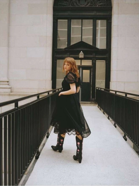 Young woman in black dress and floral boots poses on a walkway against a building with black window frames, creating a vintage aesthetic.