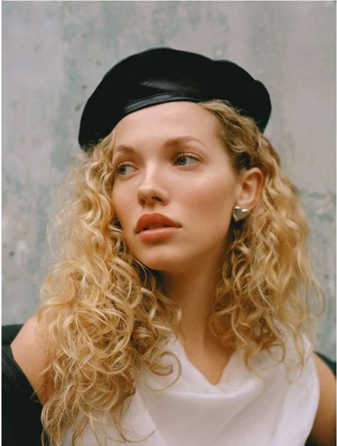 Close-up studio shot of a blonde woman wearing a black beret, white top, and silver earrings against a textured blue background.