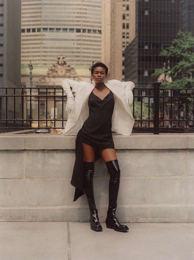Woman in black dress and glossy thigh-high boots poses against urban backdrop, wearing a white blazer with city buildings in the background.