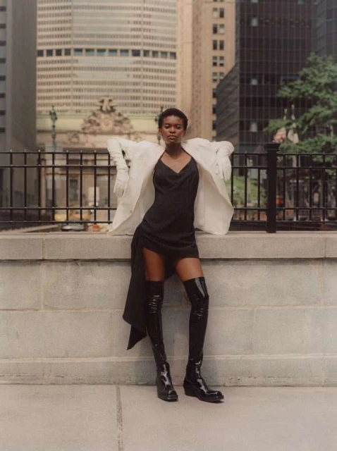 Woman in black dress and glossy thigh-high boots poses against urban backdrop, wearing a white blazer with city buildings in the background.