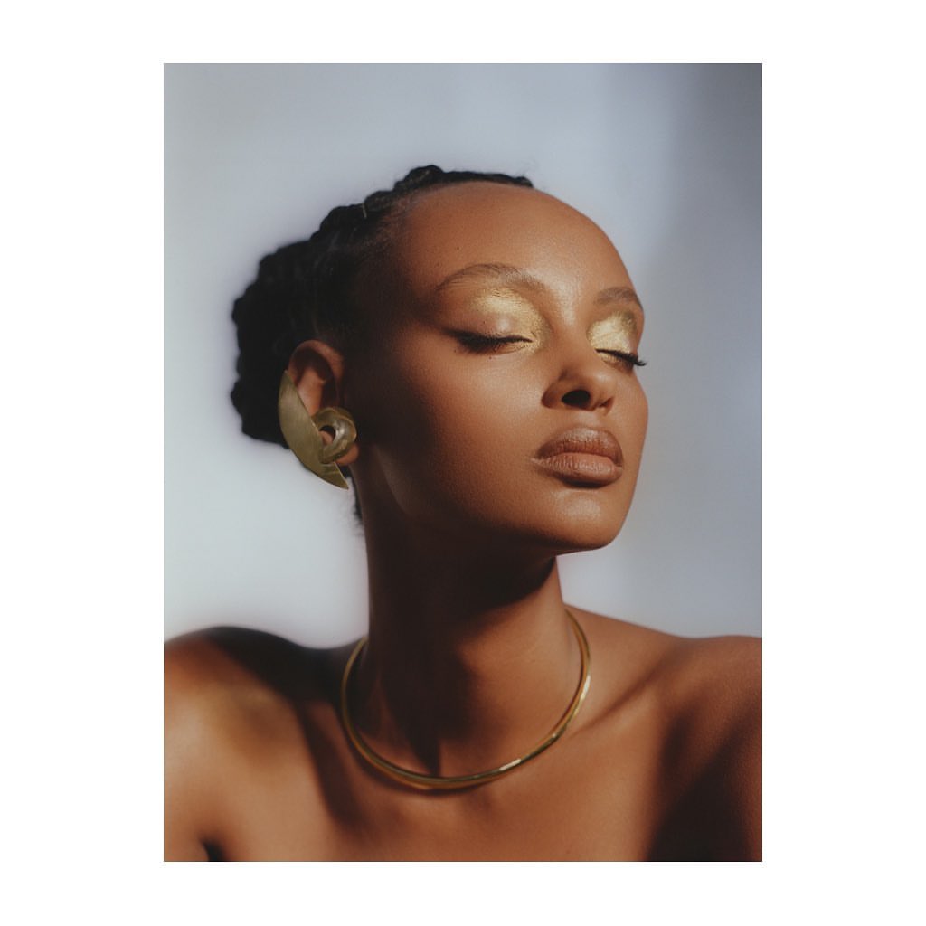 Close-up of a Black woman with elegant golden eyeshadow, a gold choker necklace, and a large gold earring, eyes closed. 