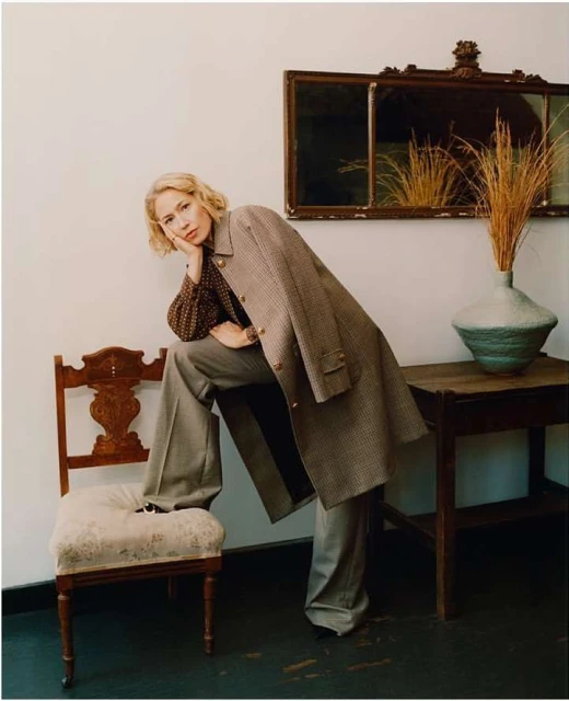 A woman in a brown patterned top and grey suit poses seated on a chair in front of a wall with mirror and vase of reeds.