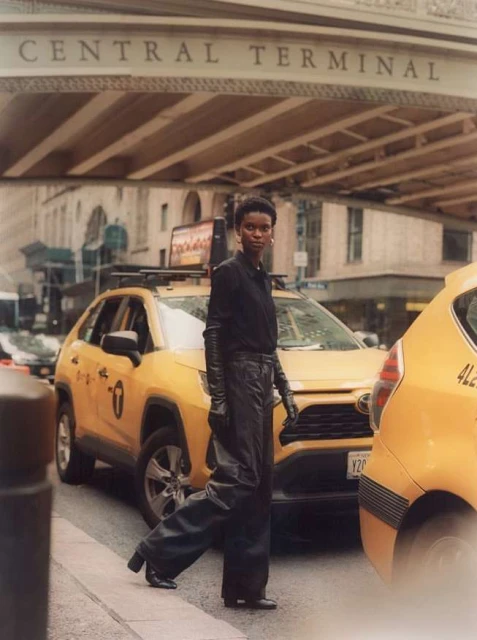 A stylish Black woman in a black outfit and gloves confidently walks across a crosswalk in front of yellow taxis and Grand Central Terminal.