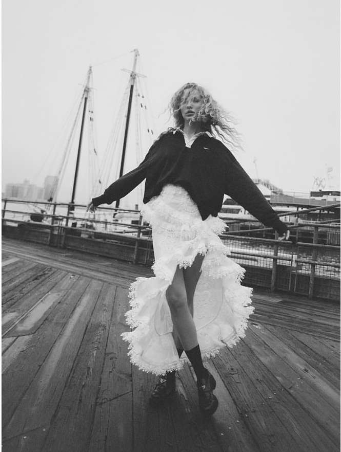 Fashion editorial shot of a blonde woman wearing a black sweater and white lace skirt posing on a wooden pier with a tall ship in the background.