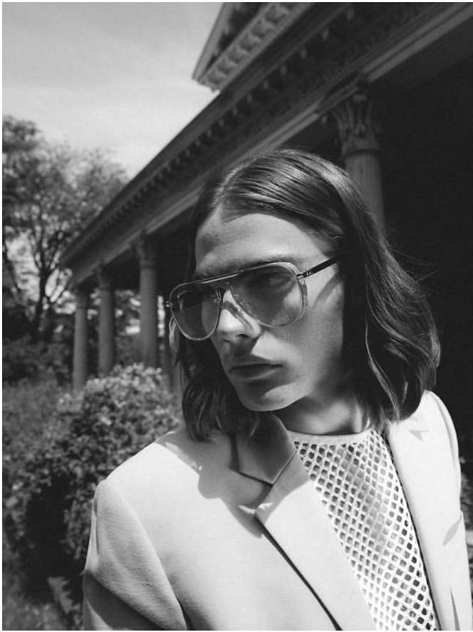 Monochrome portrait of a man with long hair wearing sunglasses, a white mesh top, and a blazer, standing in front of a colonnade.